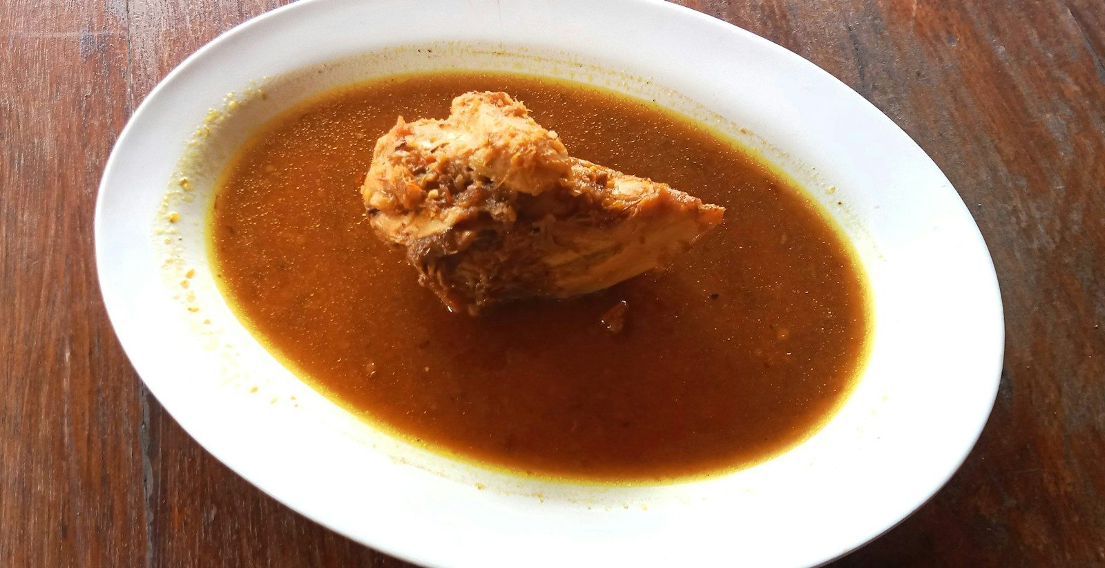 a piece of chicken in a thin red-brown soup on a white plate