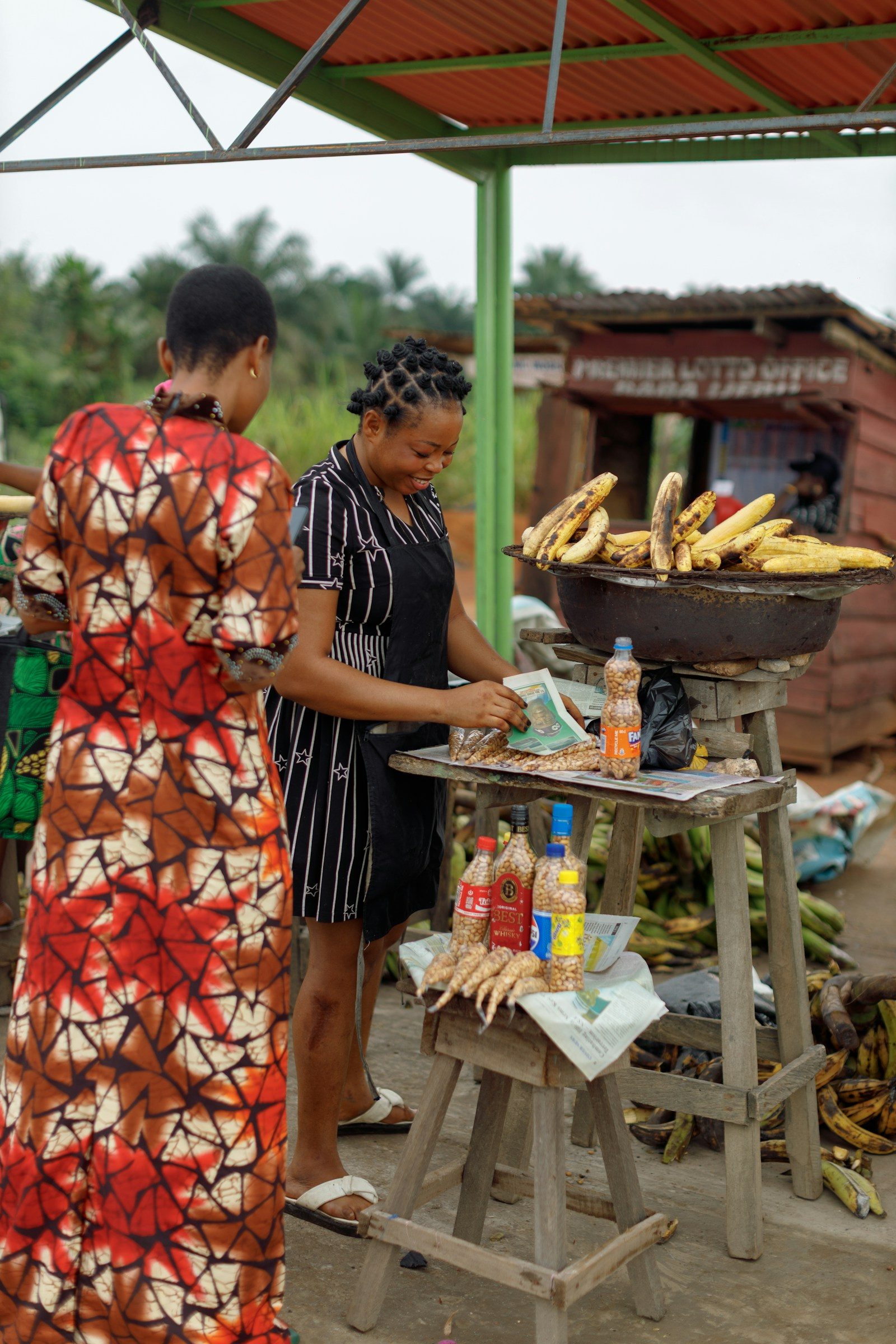 a woman selling roasted plantain and groundnuts at a roadside stall, with more plantains on the ground